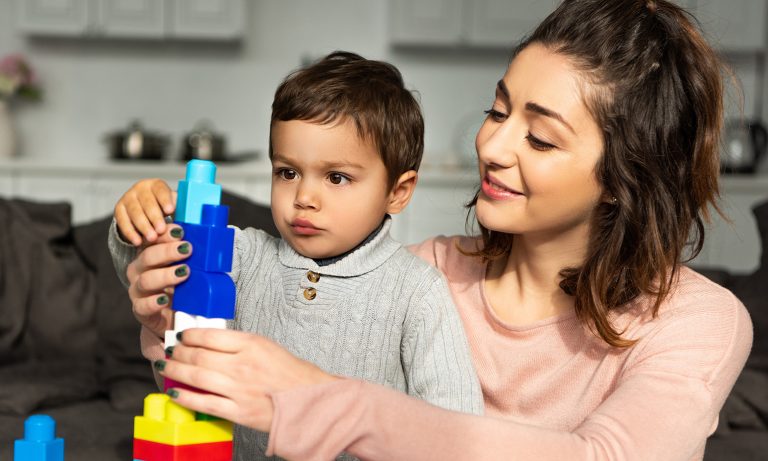 Besišypsanti mama ir mažasis sūnelis namuose žaidžia su spalvotomis plastikinėmis kaladėlėmis