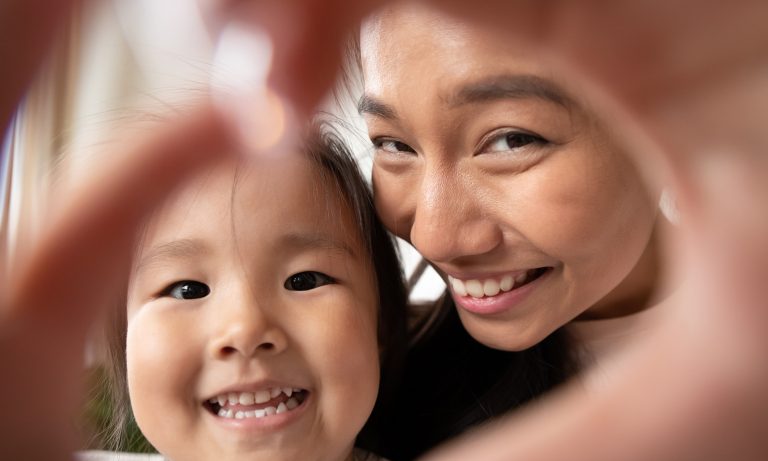 Closeup of young daughter in young mother's lap, faces together, smiling as they combine their fingers to form a heart sign