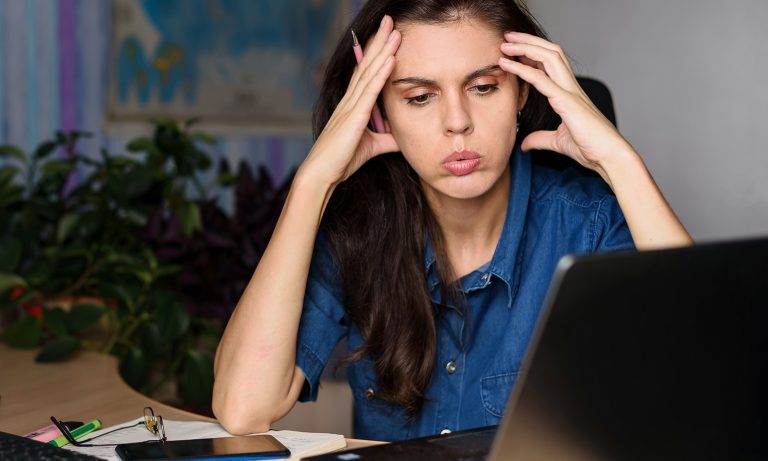 Jauna moteris prie rašomojo stalo žiūri į nešiojamąjį kompiuterį, pirštai link smilkinių, nykščiai prie žandikaulio, burna suraukta iškvepiant, patiria stresą