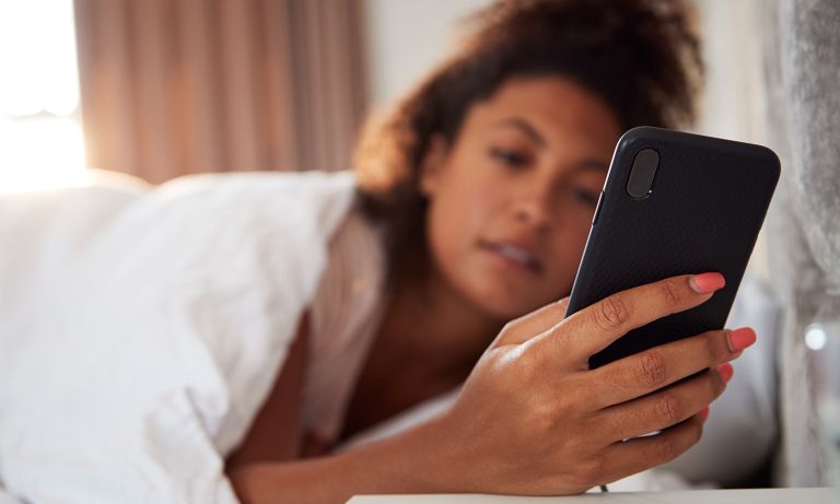 Jauna moteris guli guli lovoje, ant jos paklodė, pakreipia galvą į šoną, kad patikrintų savo mobilųjį telefoną