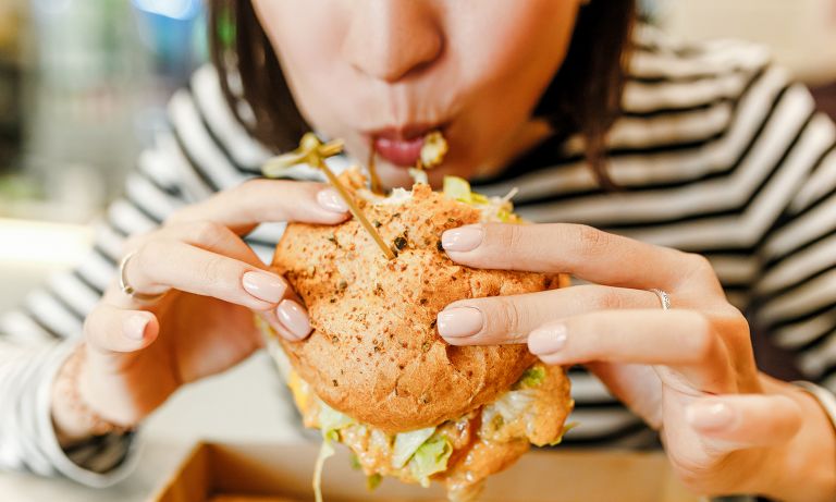 Young woman eating fast food chicken sandwich, food pick skewer sticking out of it, flecks of food hanging from her lips