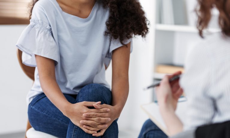 Молодая женщина, сложив руки на коленях, от голеней до плеч в кадре, консультируется с терапевтом