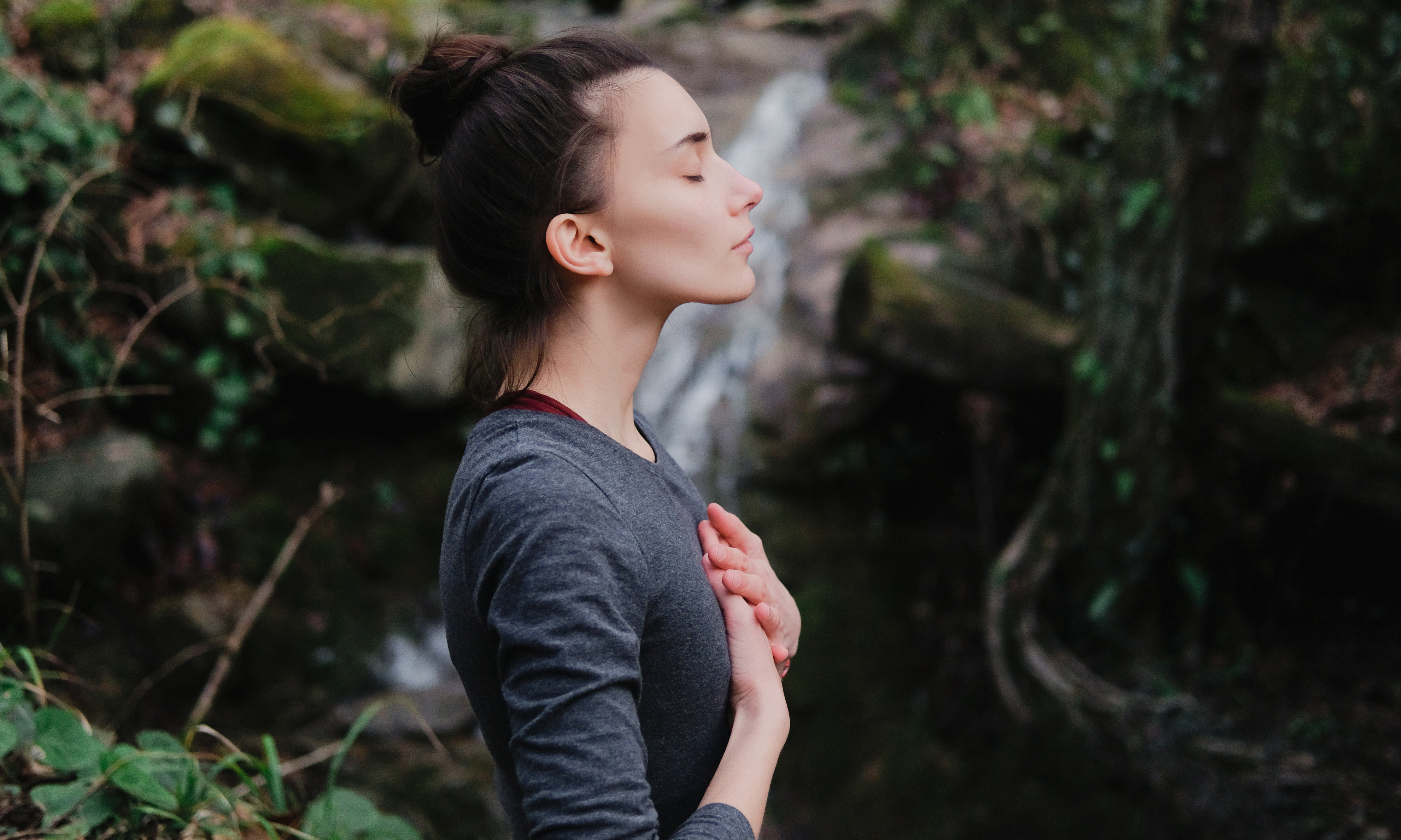 Young female yoga practitioner, hands overlapped over chest, engages in Pranayama breathing exercise outside, a waterfall behind