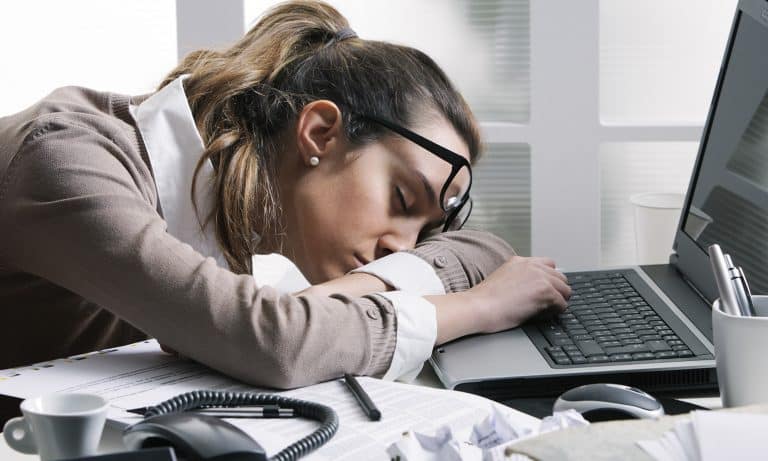 Young woman in office, asleep at laptop, chin resting on desk, eyeglasses askew