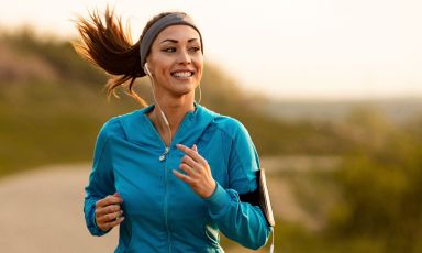 Young woman in sweat jacket smiles as she runs along dirt road, mobile music player strapped to arm