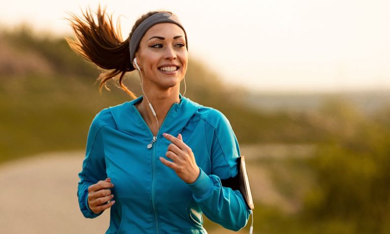 Young woman in sweat jacket smiles as she runs along dirt road, mobile music player strapped to arm