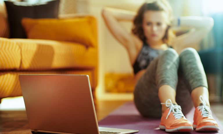 Young woman in workout clothes performs situps on yoga mat in living room while looking at laptop monitor beside her feet</p><p>